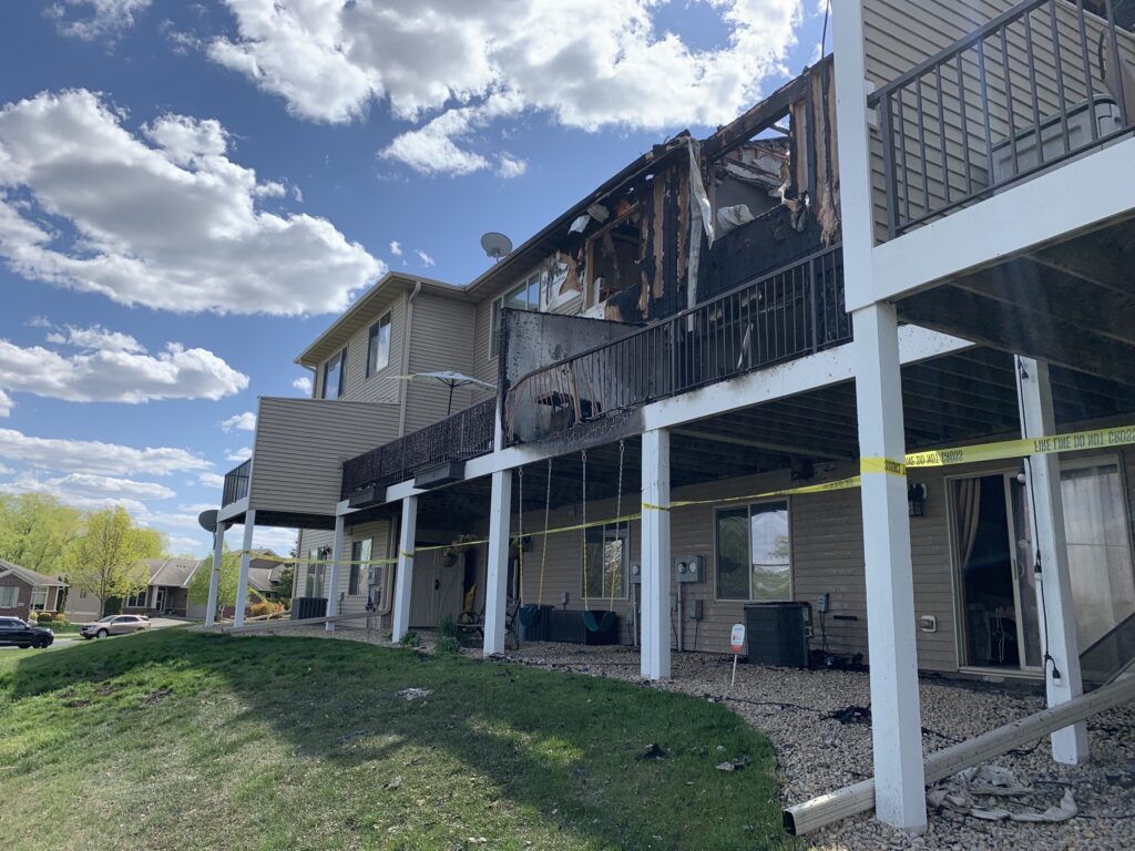 A two-story townhome with significant fire damage on an upper floor patio. Yellow caution tape is present, and the sky is partly cloudy, signaling the need for immediate fire restoration efforts.