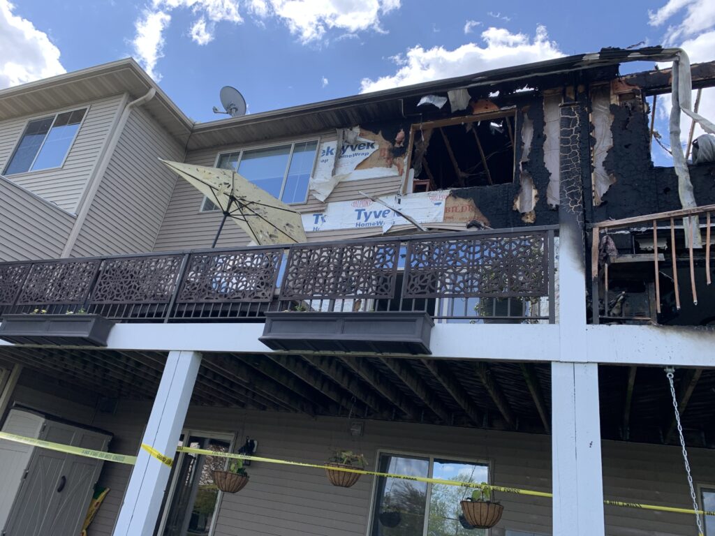 A two-story townhome with noticeable fire damage on the upper floor and charring on the exterior. The balcony railing is intact, with an umbrella and some plants visible. Yellow caution tape is in place, pending fire restoration efforts.