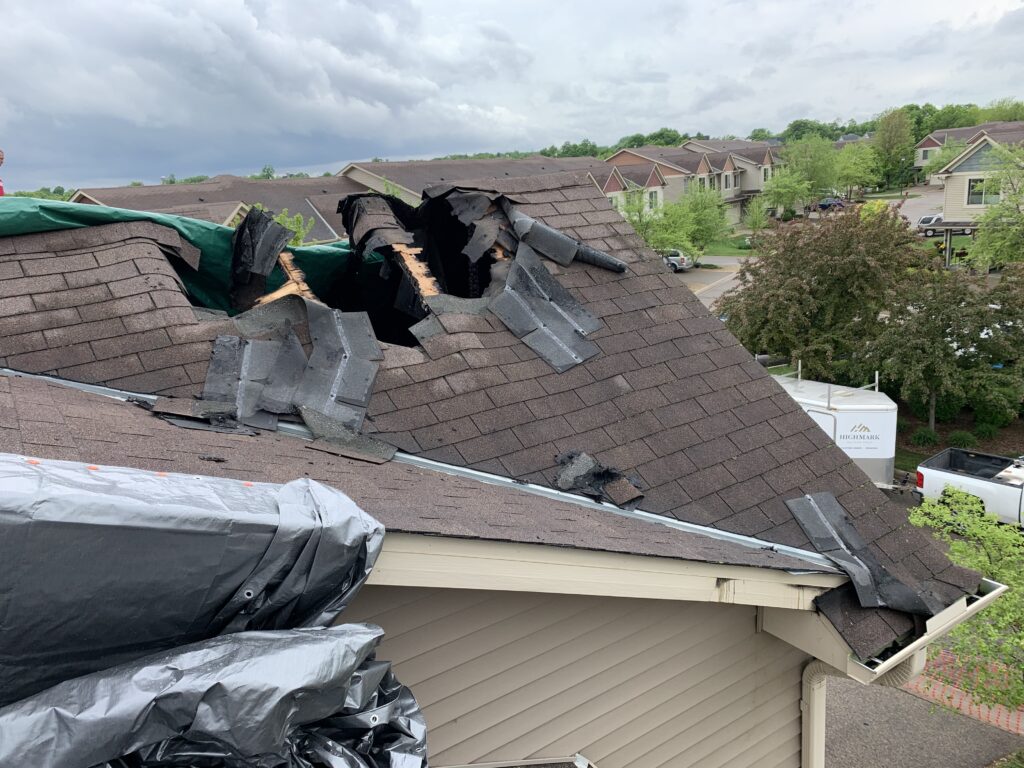 A roof with significant damage, including large holes and missing shingles, likely caused by a storm. The surrounding area includes residential buildings and a cloudy sky, necessitating immediate townhome fire restoration to ensure the safety and integrity of the neighborhood.