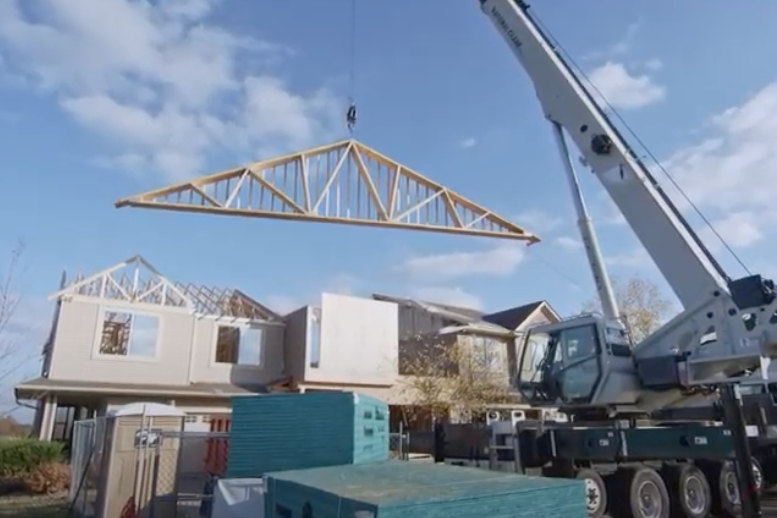 A crane hoists a large wooden roof truss at a residential construction site. Partially built houses, likely part of an ongoing townhome fire restoration, and stacks of green construction materials are visible in the background.