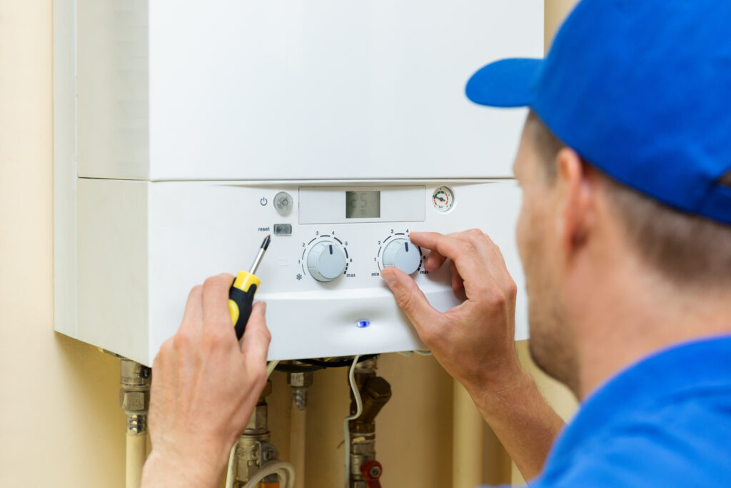 A technician wearing a blue cap and holding a screwdriver adjusts the dials on a white wall-mounted boiler, ensuring it operates efficiently to prevent frozen and burst pipes during winter.