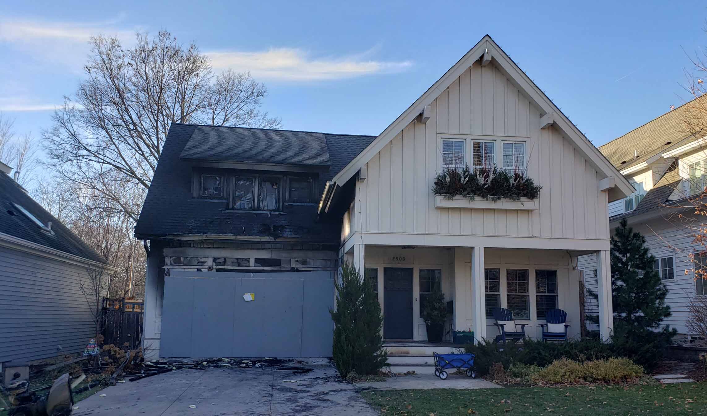 Two-story house on Upton Avenue with visible fire damage on the garage's exterior and scorched roof shingles. The rest of the house appears intact with a small front porch and potted plants, making it a prime candidate for a thoughtful remodel.