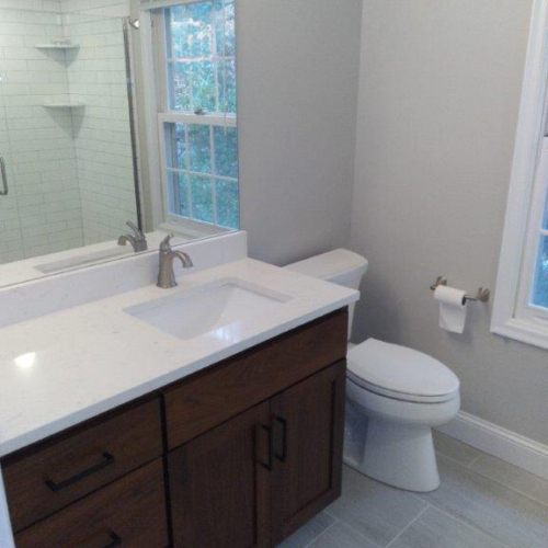 A beautifully remodeled bathroom in the Apple Valley house features a wooden vanity, white countertop, sink, toilet, and large mirror. In the background, a window and glass-enclosed shower add to the elegant design.