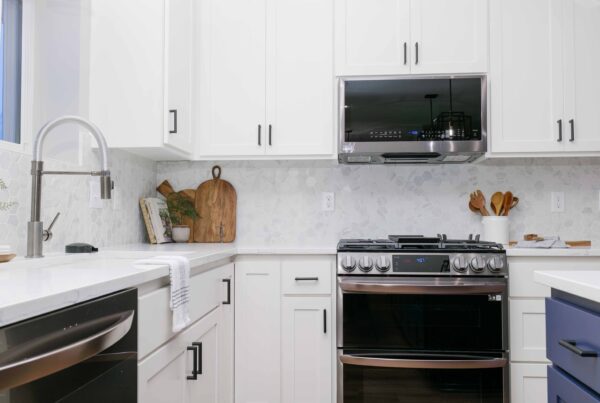 A modern Lakeville Kitchen Fire setting features white cabinets, stainless steel appliances, a cutting board set against the backsplash, a gas stove, a large faucet, and a white countertop with a cloth draped over the sink.