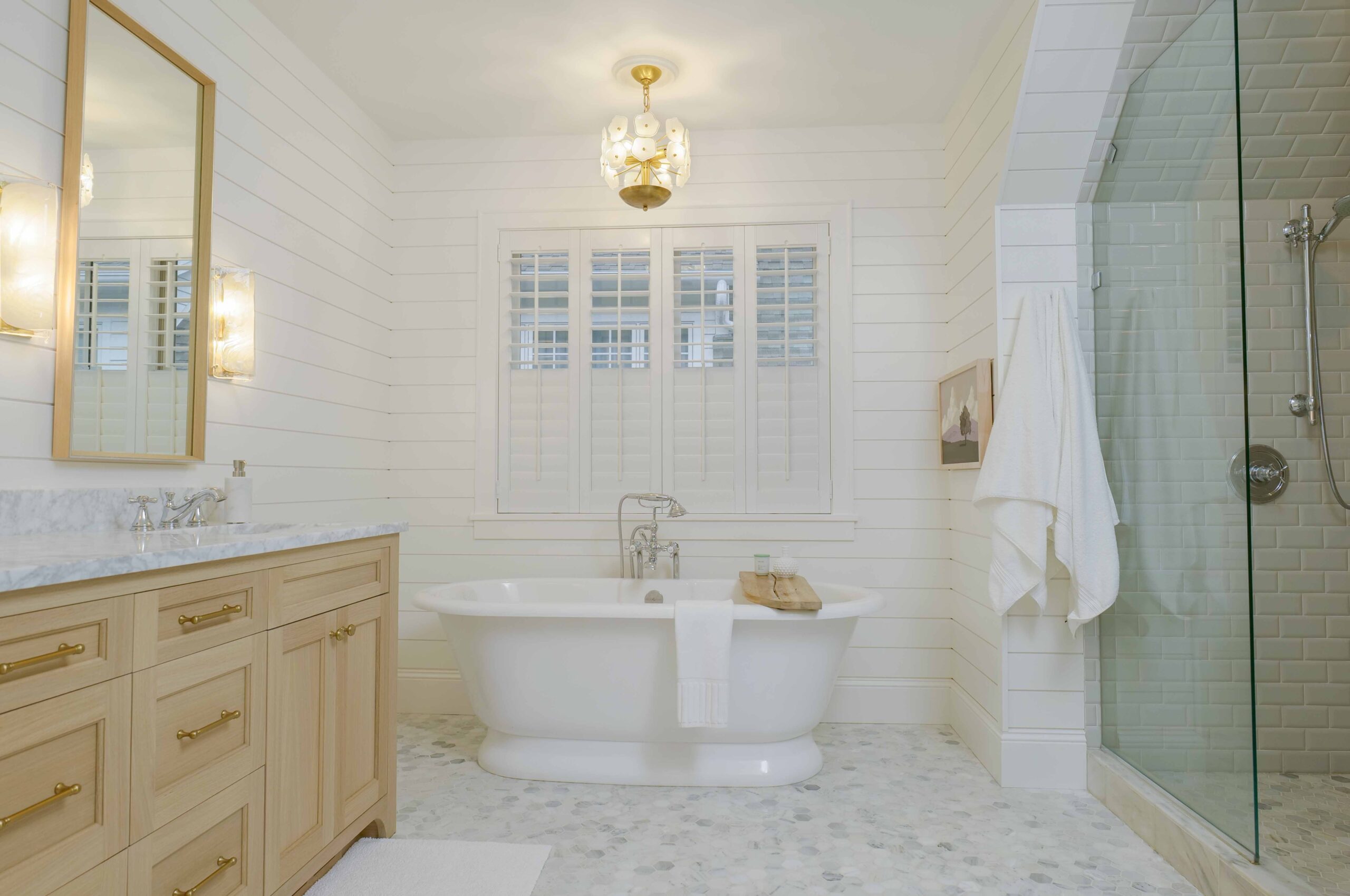 A light and airy bathroom, part of the Upton Avenue Fire Remodel, featuring a wooden vanity, a freestanding bathtub, a glass-enclosed shower, white shiplap walls, and a hanging light fixture.