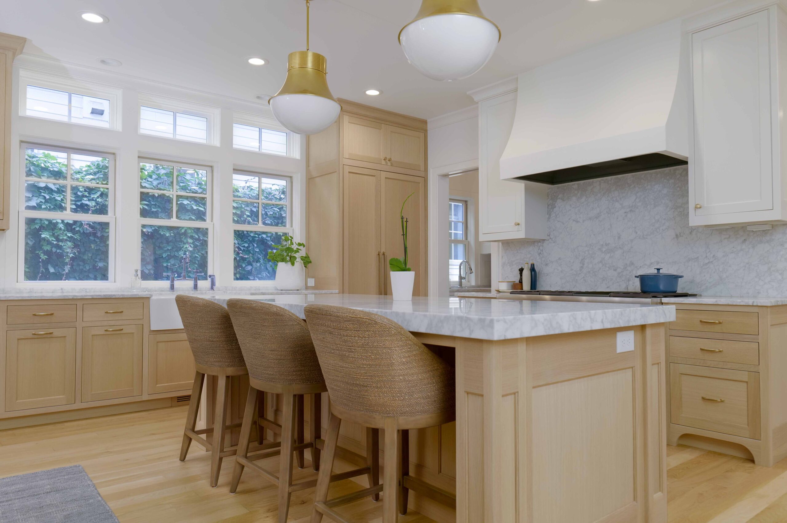 A modern kitchen with light wood cabinets, a marble countertop island with three bar stools, pendant lights, a white range hood, and large windows offering natural light is part of the incredible Upton Avenue Fire Remodel.