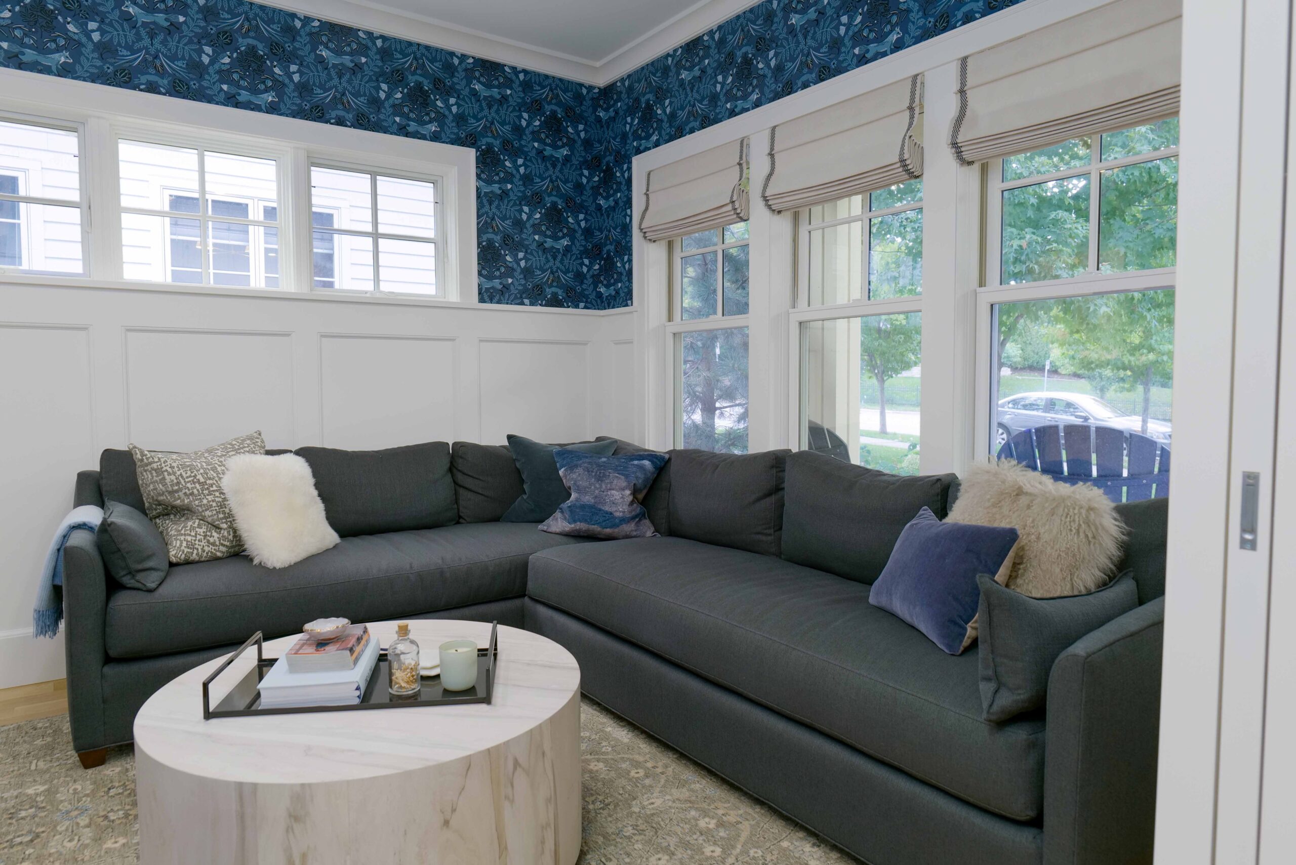 A living room with a dark gray sectional sofa, various throw pillows, a round marble coffee table, and blue patterned wallpaper above white wainscoting. The Upton Avenue Fire Remodel boasts large windows with Roman shades in the background.