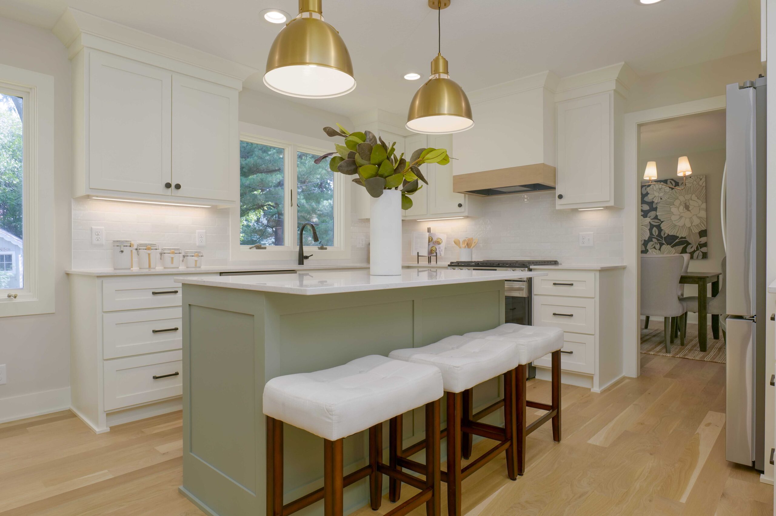 A modern kitchen with white cabinetry, a central island with stools, large gold pendant lights, and a view into a dining area with patterned wallpaper shines after an expert Edina water damage remodel.