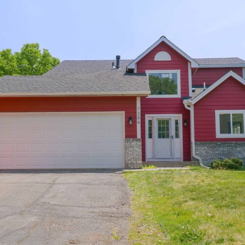 A two-story house in Apple Valley with a red exterior, white trim, and a gray shingled roof. It has a large white garage door and a front yard with green grass and trees. Recently remodeled after a house fire, it offers fresh interiors that blend modern comfort with classic charm.